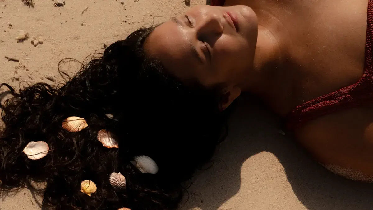 Person resting on beach sand, curly hair fanned out and dotted with seashells.