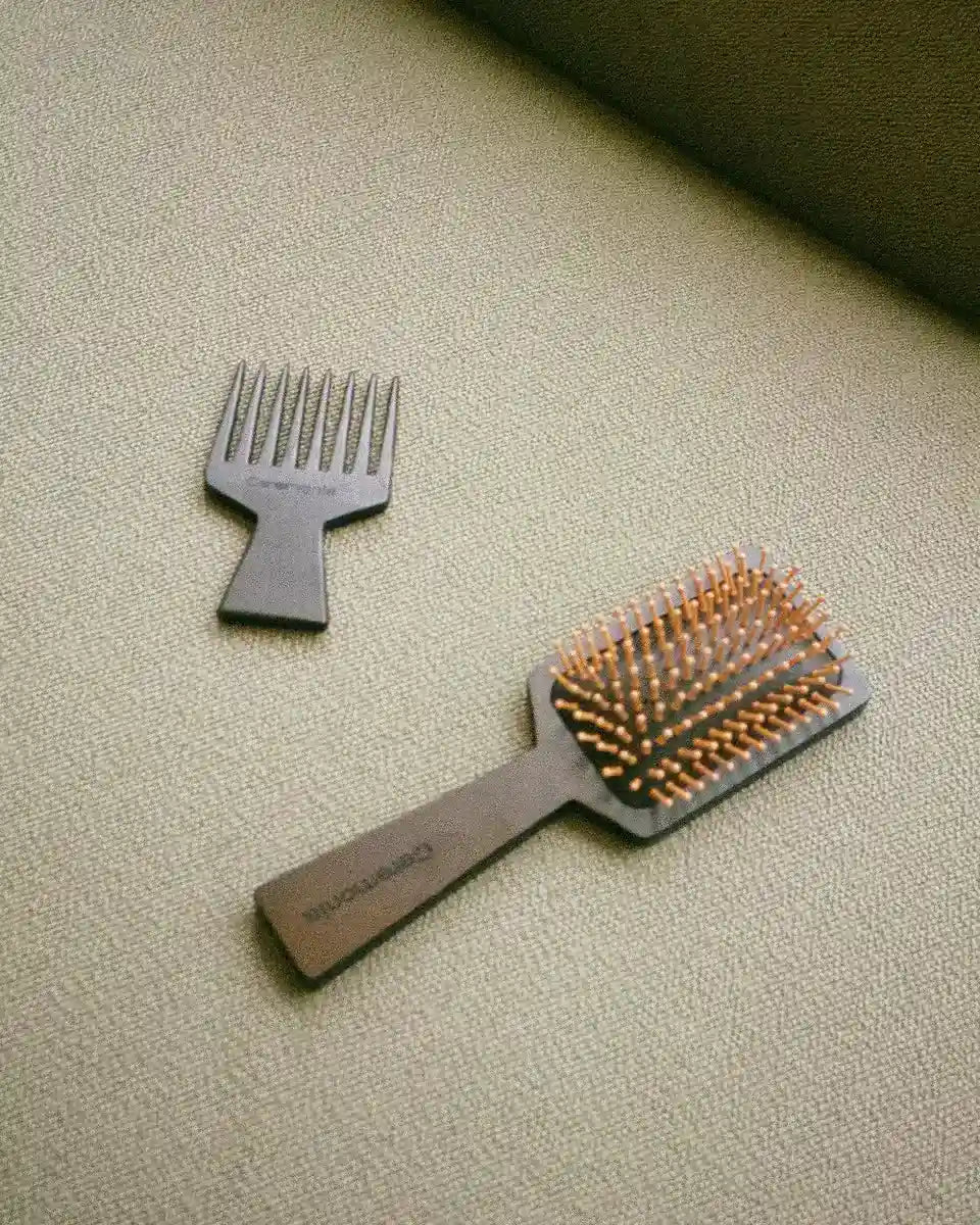 Hair brush with wooden handle and gold bristles next to a wide-tooth comb on a beige fabric surface.