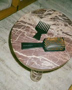 Hair brush with gold bristles and wide-tooth comb on a round marble table with a plush rug underneath.