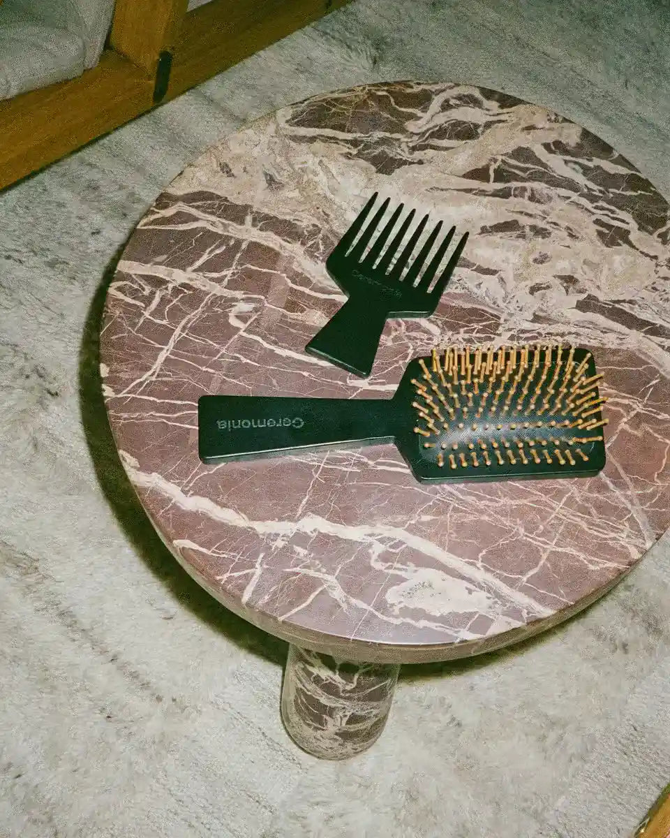Hair brush with gold bristles and wide-tooth comb on a round marble table with a plush rug underneath.