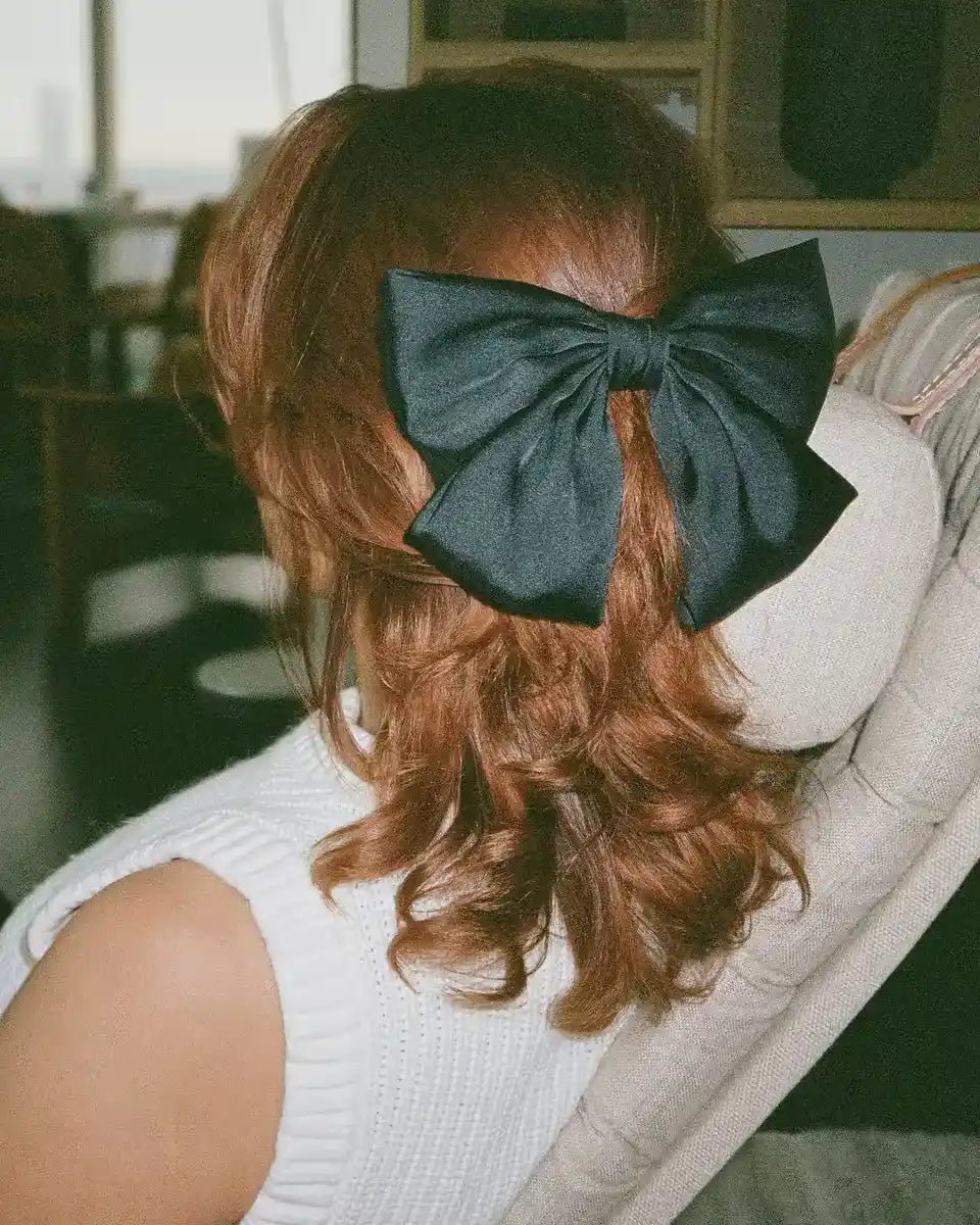 Black hair bow styled on wavy auburn hair, worn with a white sleeveless knit top indoors.