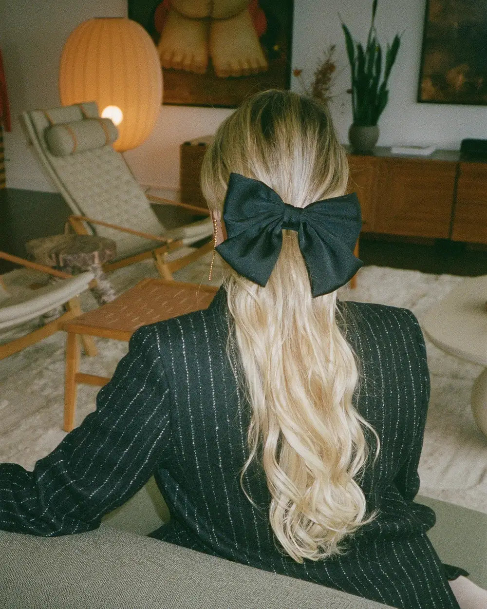 Black hair bow styled in long wavy blonde hair, viewed from behind in a cozy interior setting.