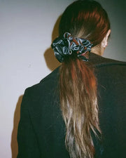 Black scrunchie securing a low ponytail on long brown hair, worn with a dark coat against a plain background.