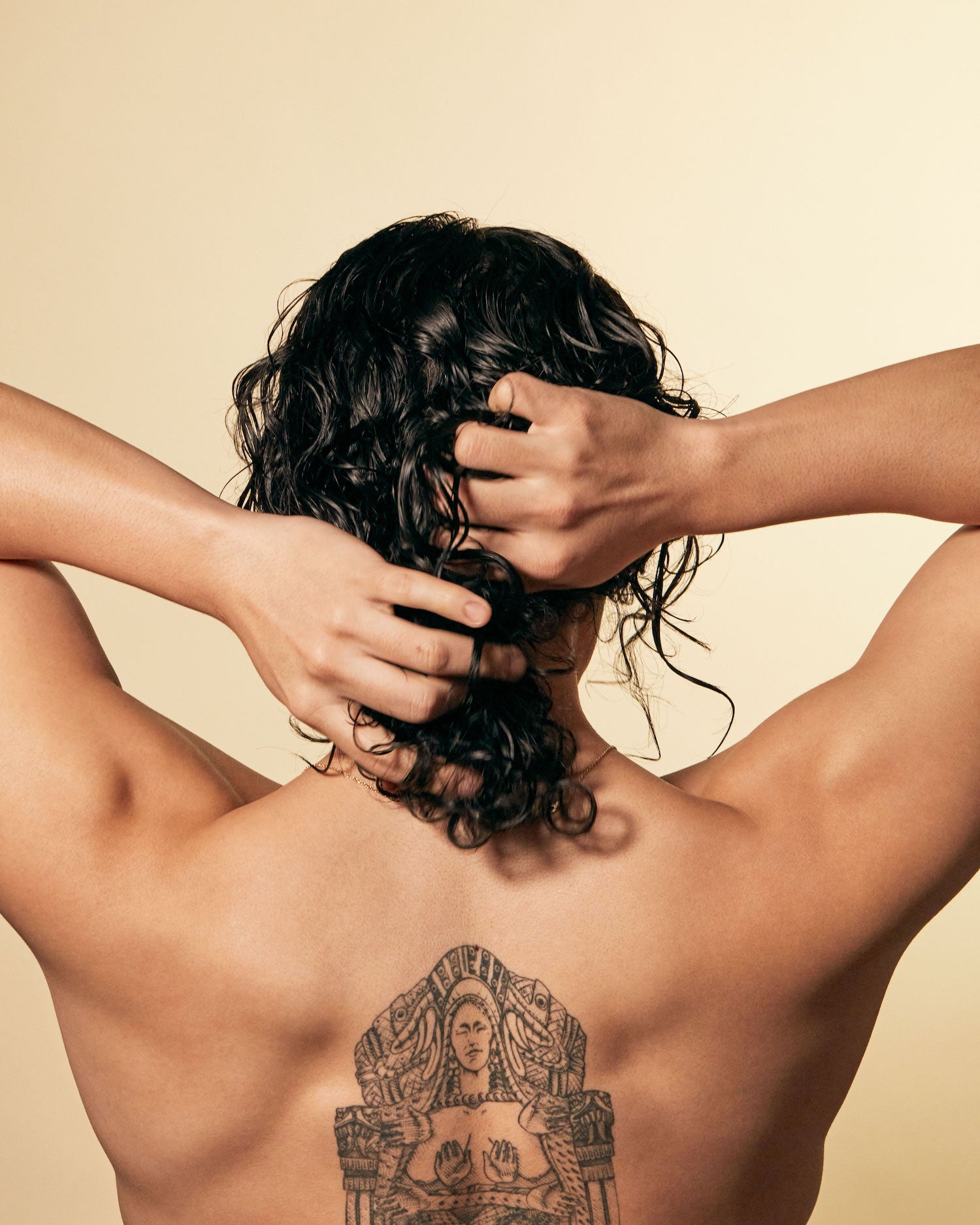Leave-in conditioner being applied to wet, curly hair by a person with a large back tattoo against a neutral background.