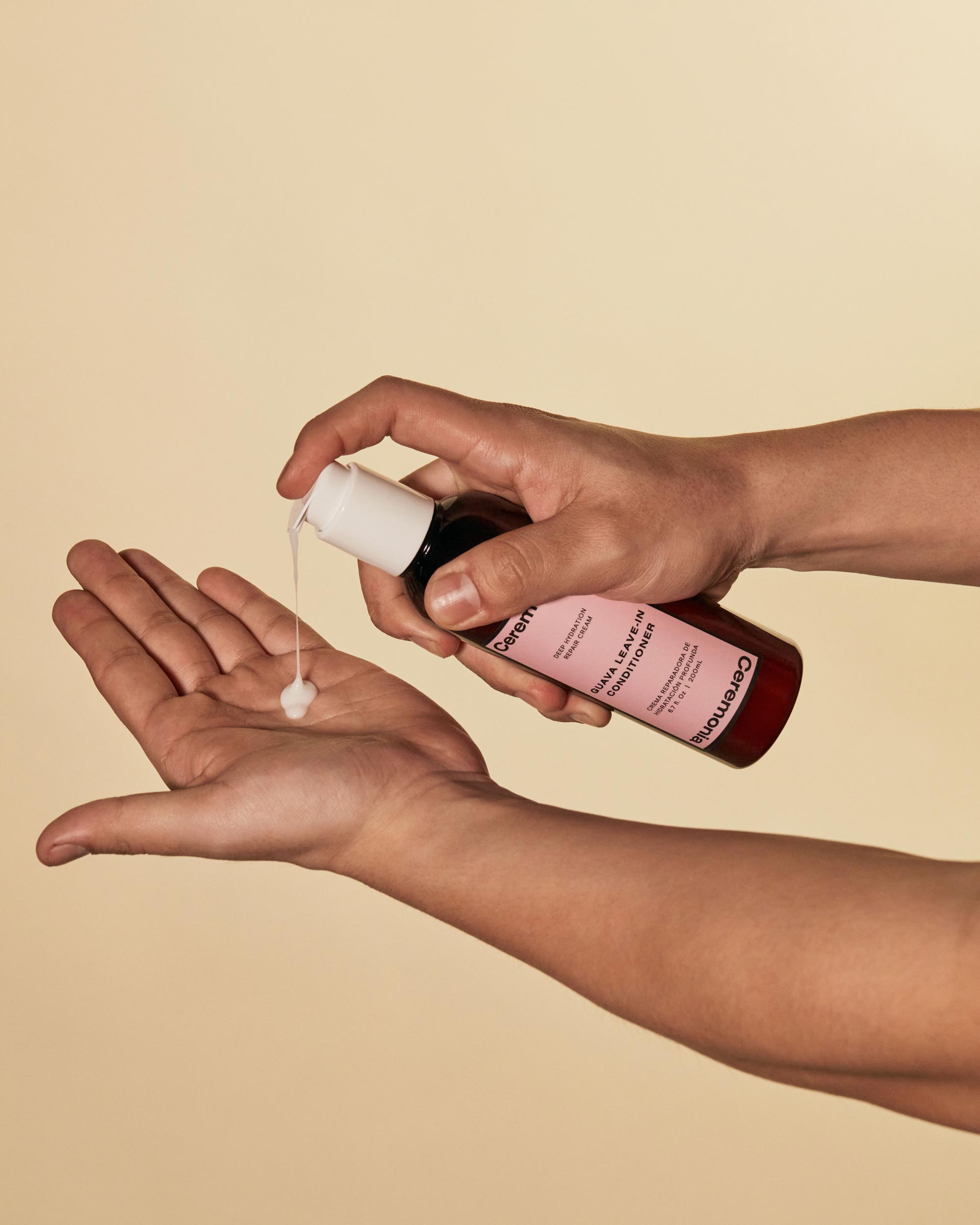 Hair repair cream being dispensed from a Ceremonia Guava Leave-In Conditioner bottle into an open palm against a beige background.