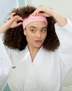 Towel headband in pink worn by a woman with curly hair while getting ready in a white waffle-textured robe.