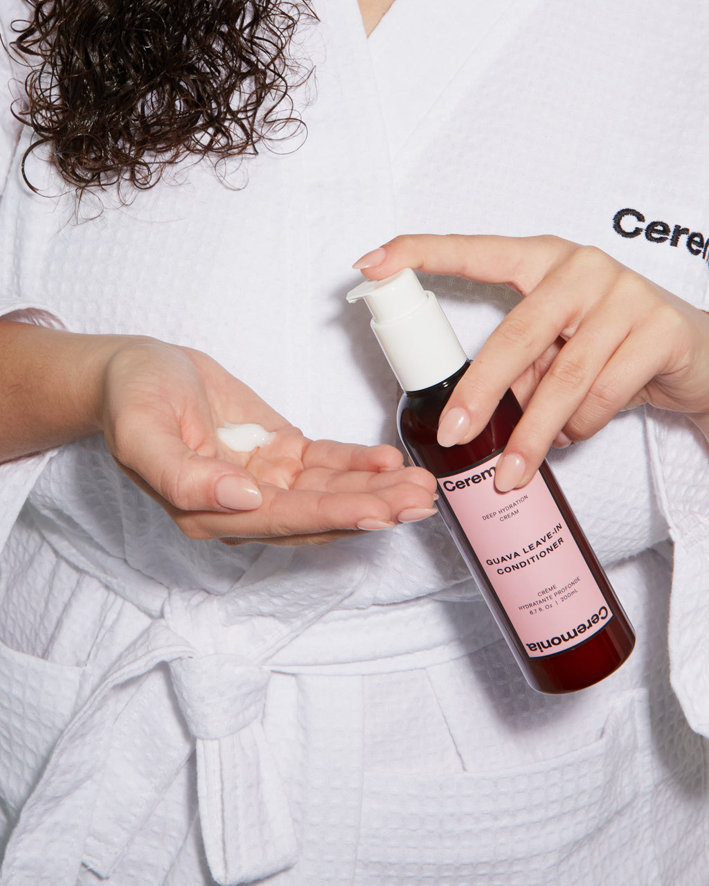 Guava leave-in conditioner being dispensed into a woman's hand while she wears a white robe with damp curly hair.