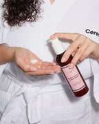 Guava leave-in conditioner being dispensed into a woman's hand while she wears a white robe with damp curly hair.