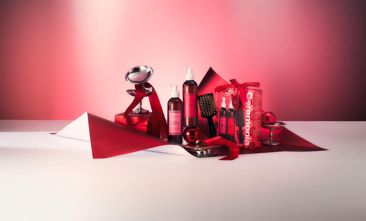 A curated collection of red-themed beauty and grooming products arranged on a white surface with dramatic red and pink lighting. Visible items include several bottles, a small mirror, a black comb, and a red gift box with white lettering, all styled with red ribbon and paper accents.
