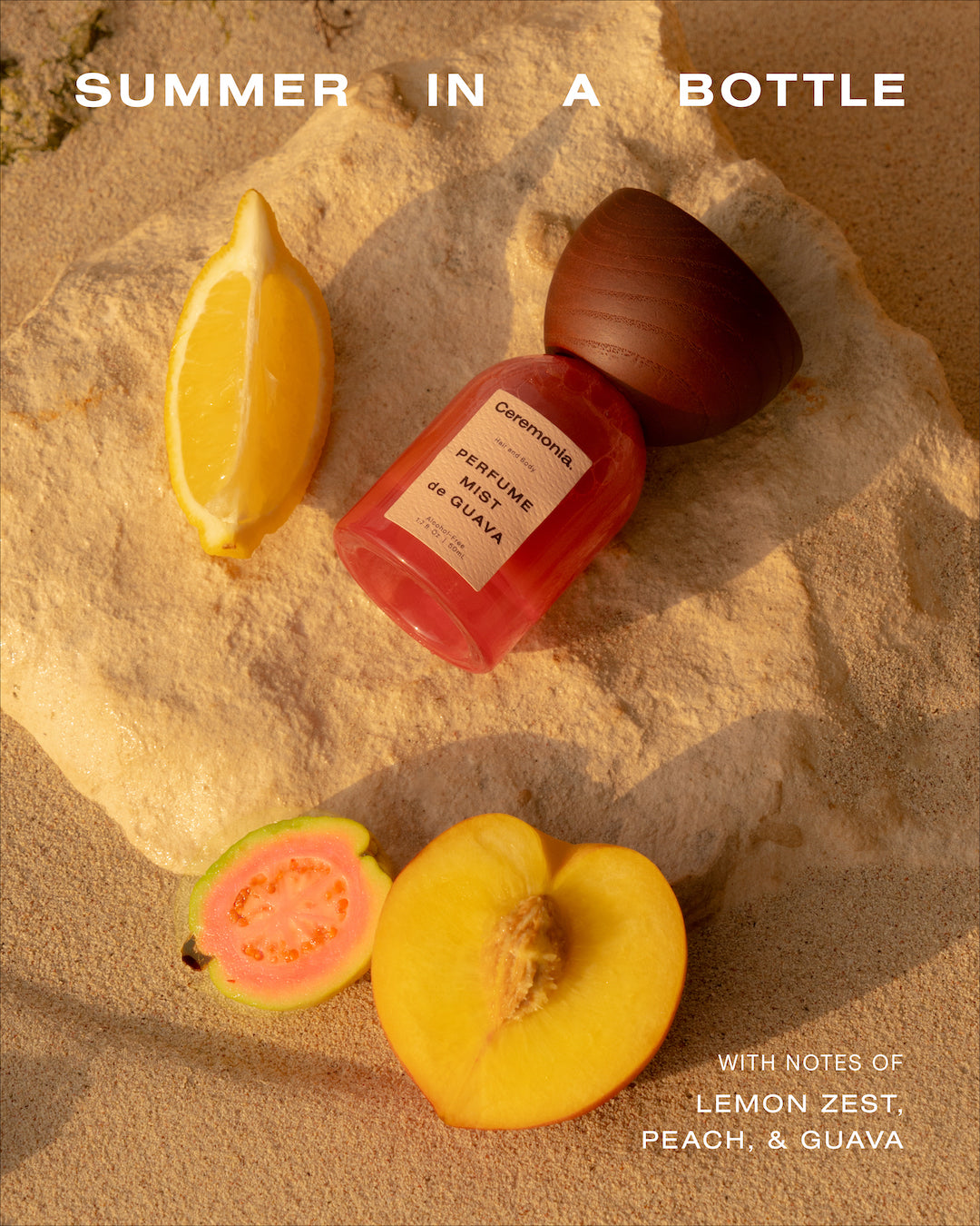 Guava perfume mist bottle on sand beside lemon, peach, and guava slices, described as summer in a bottle.