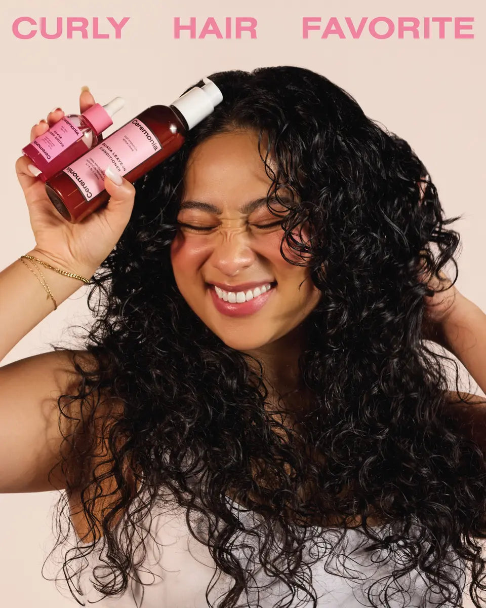 Smiling person with curly hair holding Ceremonia Guava Gloss Drops and leave-in conditioner duo.