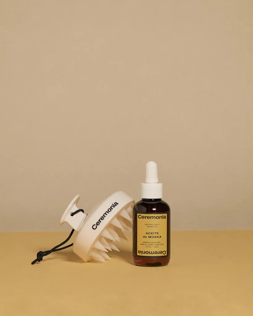 Ceremonia Aceite de Moska scalp oil dropper bottle beside a white silicone scalp massager on a tan backdrop.