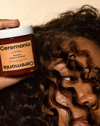 Ceremonia Papaya Scalp Scrub jar held beside curly hair in a warm-lit close-up portrait.