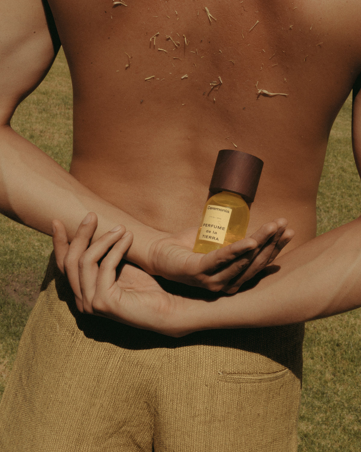 Citrus perfume mist de la Tierra held behind the back of a shirtless person standing on sunlit grass.