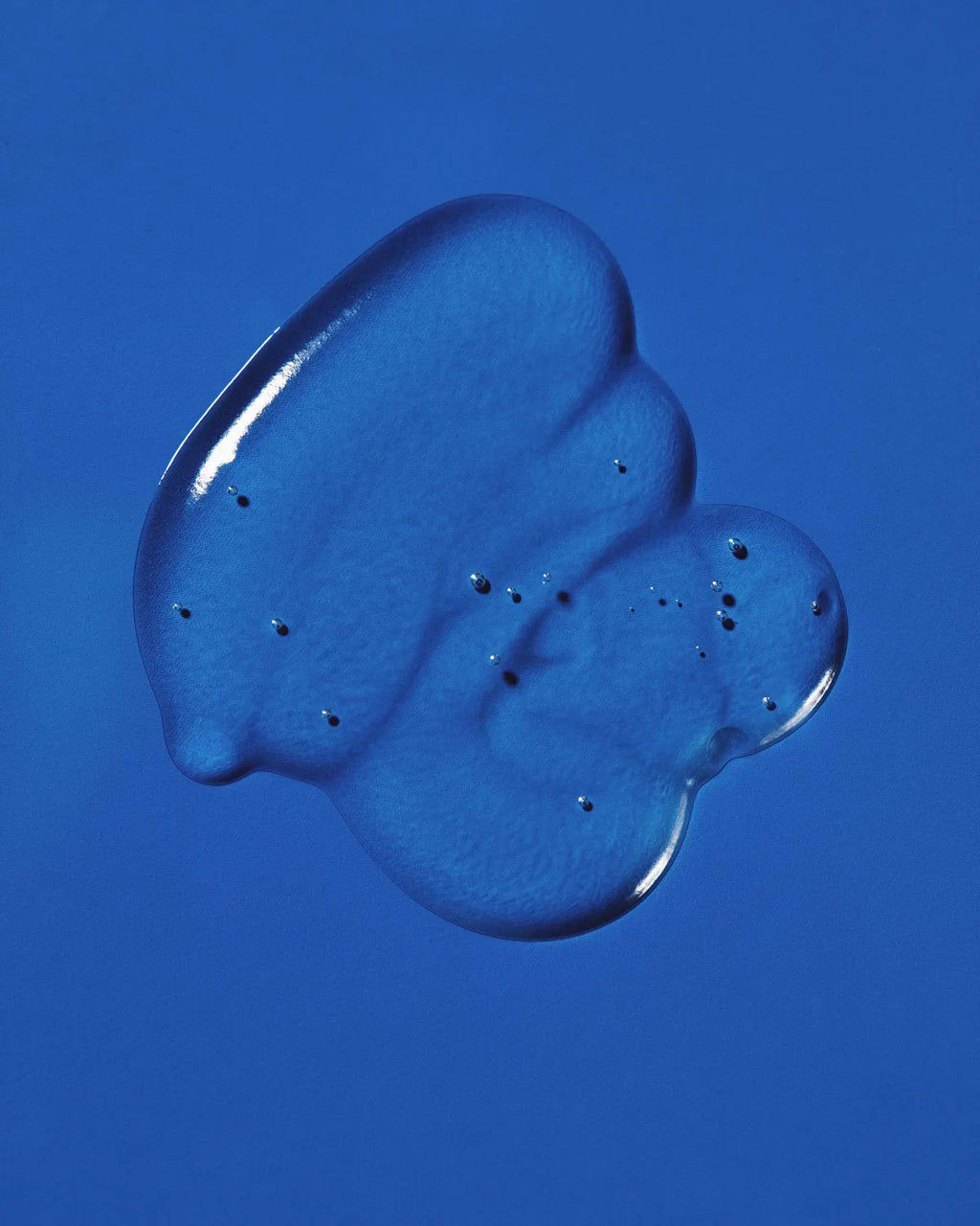 Clear dollop of Pequi styling gel swatched on a bright blue background, showing its glossy, bubble-flecked texture.