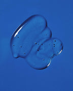 Clear dollop of Pequi styling gel swatched on a bright blue background, showing its glossy, bubble-flecked texture.