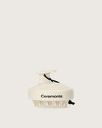 Cream-colored Ceremonia scalp massager with silicone bristles and a black wrist strap.