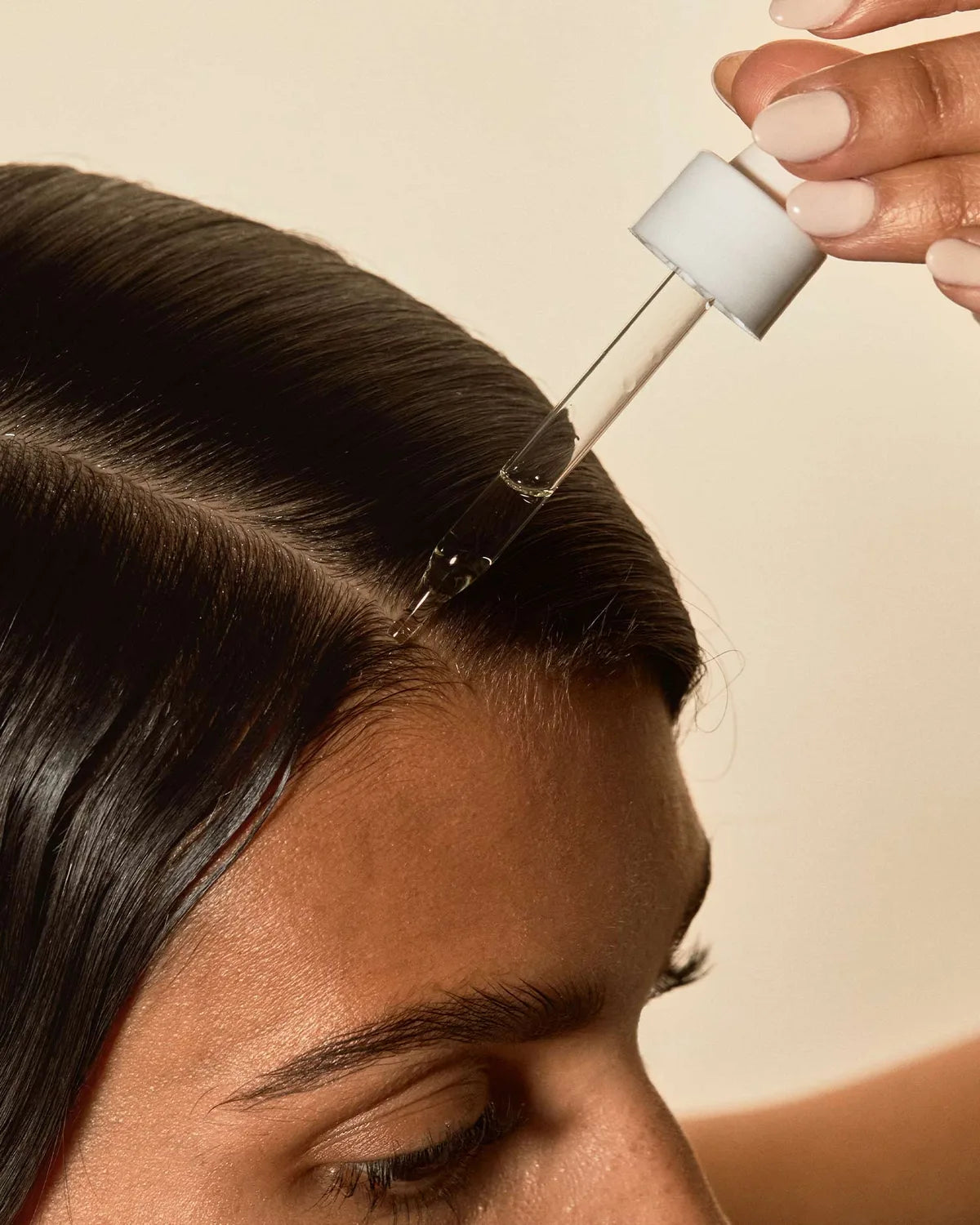 Dropper applying Aceite de Moska scalp oil along a clean center part on dark hair.