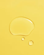 Golden droplet of Aceite de Moska scalp oil pooled on a pale yellow surface.