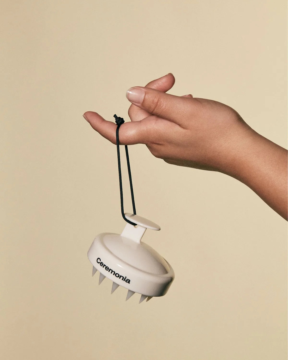 Hand dangles cream Ceremonia Scalp Masajeador by its black cord against a beige backdrop.