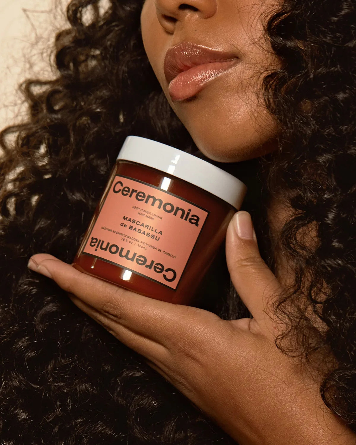 Hand holding a Ceremonia Mascarilla de Babassu deep conditioning hair mask jar beside curly hair.
