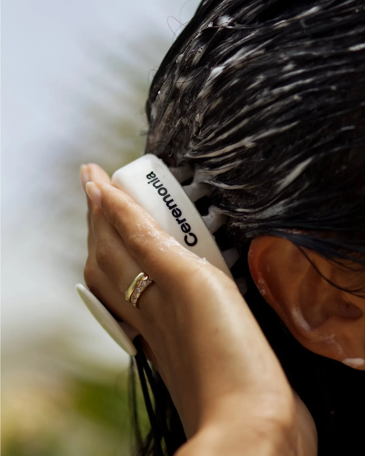 Hand using Ceremonia scalp masajeador brush to massage lather into wet dark hair.