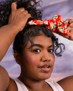 Hands tying curly hair with a red guava-print satin Ceremonia scrunchie.