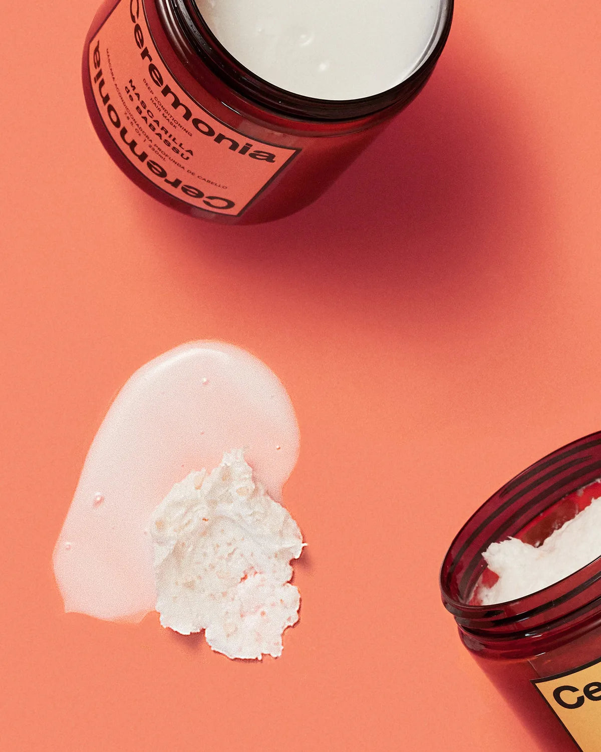 Open jars of Ceremonia Mascarilla de Babassu hair mask and Papaya Scalp Scrub with creamy texture swatches on a coral.