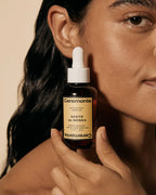 Person holding Ceremonia Aceite de Moska scalp oil dropper bottle beside their face.