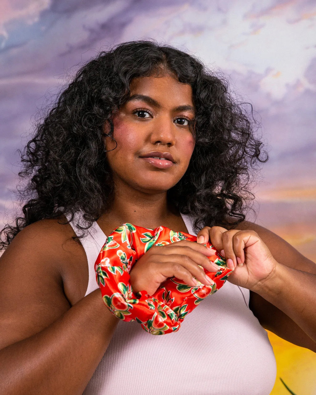 Person with curly hair holding the red guava-print Ceremonia satin scrunchie at chest level.