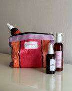 Pink and orange woven Ceremonia Pouch de Mercado holding haircare bottles on a cream surface.