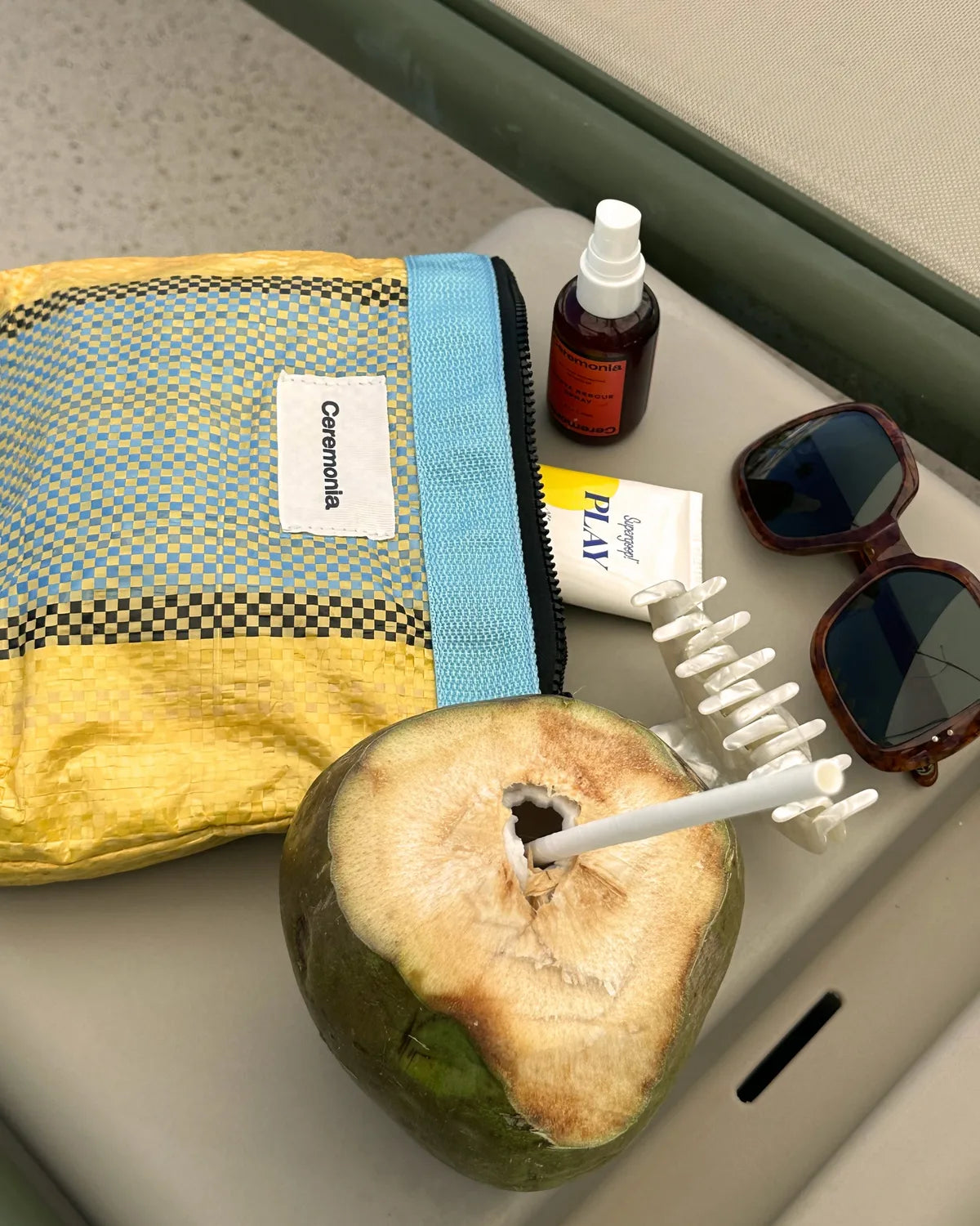 Yellow checkered Ceremonia Pouch de Mercado poolside with sunscreen, sunglasses, hair claw, and coconut.