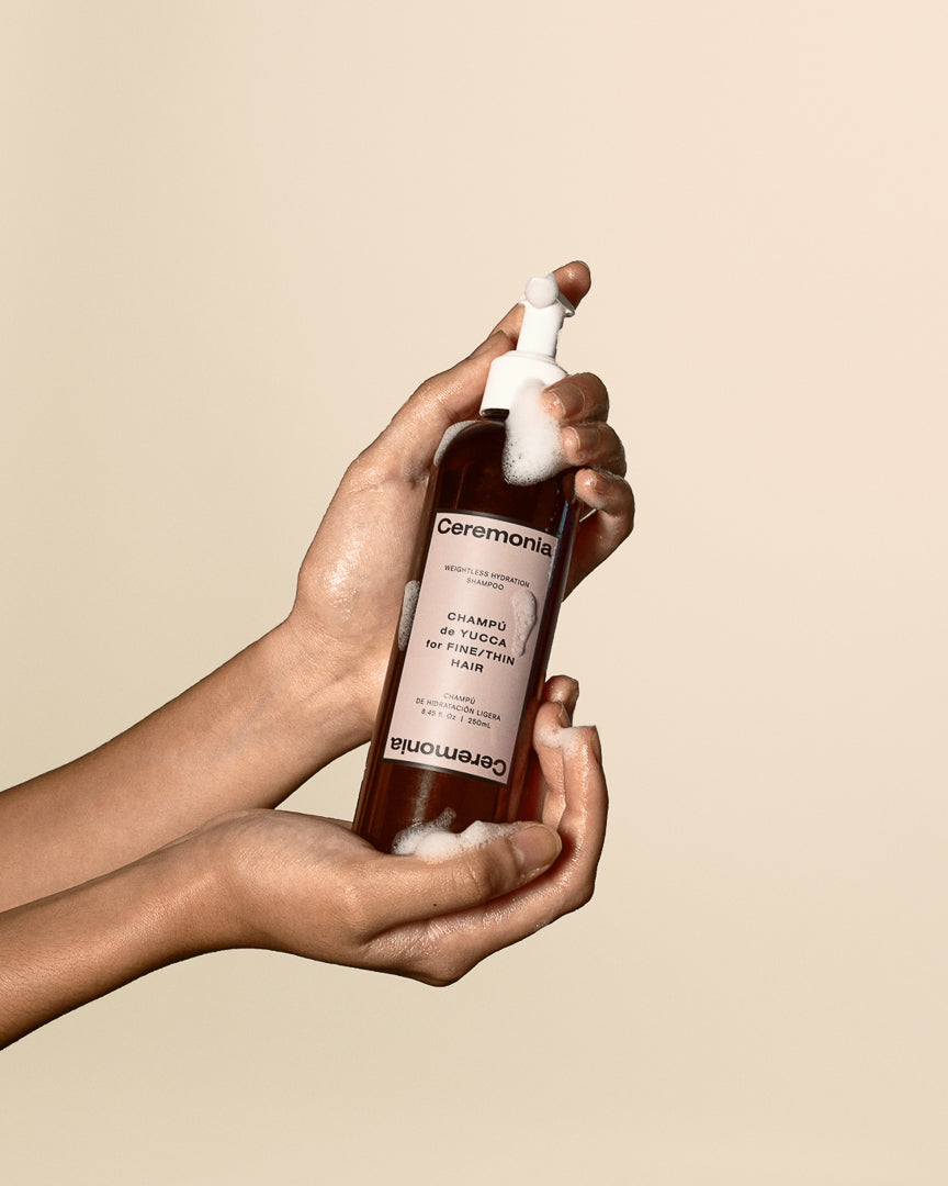 Hands holding Ceremonia weightless hydration shampoo bottle with foamy pump against a beige backdrop.